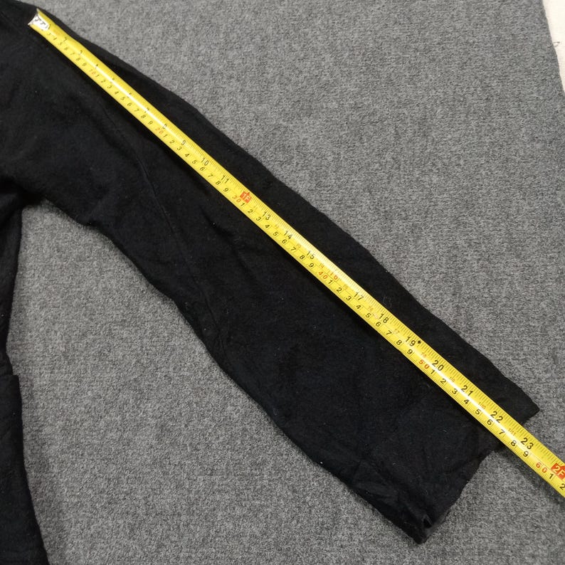May include: A black long-sleeved garment sleeve positioned next to a yellow measuring tape. The tape shows the sleeve length is approximately 23.5 inches from the shoulder seam to the cuff edge, providing a clear visual reference for the size.