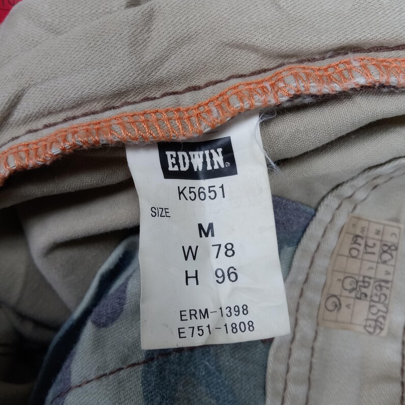 May include: A beige and orange clothing label with the text "EDWIN" and the model number "K5651". The label also includes the size "M", waist measurement "W 78", hip measurement "H 96", and the numbers "ERM-1398" and "E751-1808".