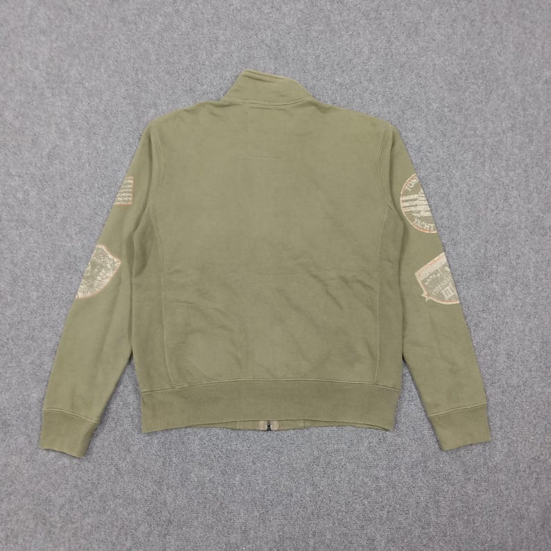 May include: Olive green zip-up jacket with a stand-up collar. The jacket features patches on the sleeves with text and graphic designs. The jacket has a full-length zipper and ribbed cuffs and hem. The jacket is made of a soft, comfortable material.