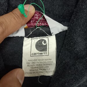 Puede incluir: Una etiqueta negra de Carhartt con texto blanco que dice "carhartt" e instrucciones para lavar la prenda: "MACHINE WASH WARM USE ONLY NON-CHLORINE BLEACH WHEN DESIRED. TUMBLE DRY LOW. REMOVE PROMPTLY STEAM IRON WHEN DESIRED."