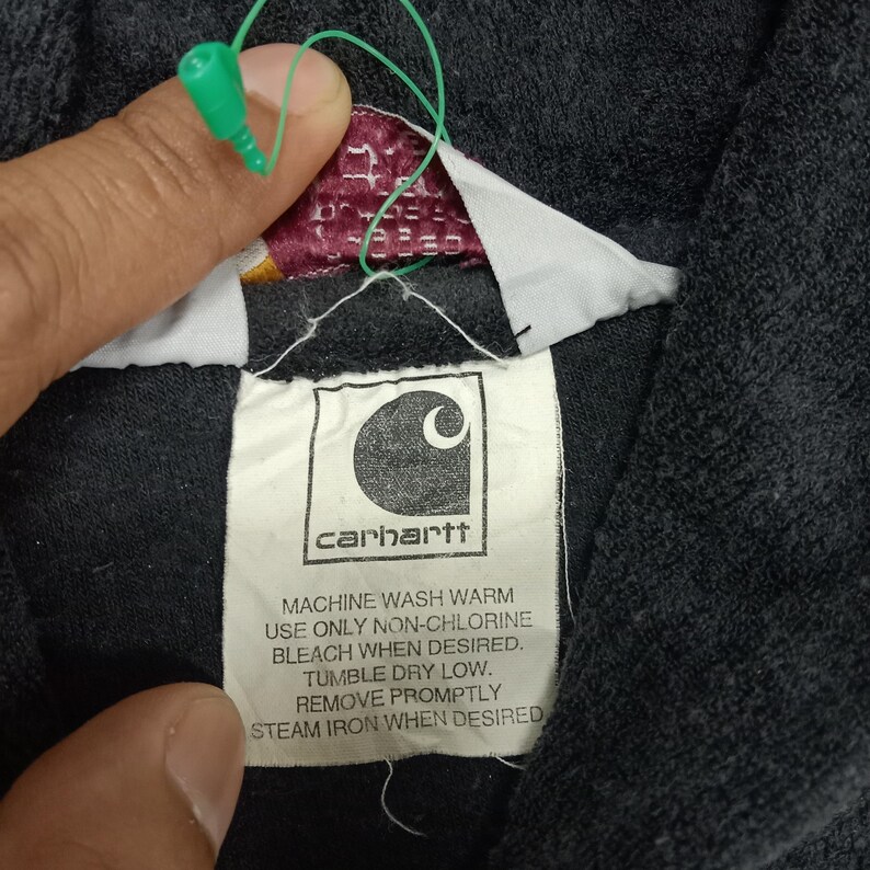 Puede incluir: Una etiqueta negra de Carhartt con texto blanco que dice "carhartt" e instrucciones para lavar la prenda: "MACHINE WASH WARM USE ONLY NON-CHLORINE BLEACH WHEN DESIRED. TUMBLE DRY LOW. REMOVE PROMPTLY STEAM IRON WHEN DESIRED."
