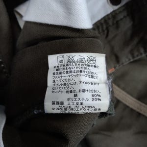 May include: A close-up of a green garment care label with washing instructions. The label is attached to a pair of green pants. The label includes a 40 degree Celsius washing symbol, a do not bleach symbol, a do not tumble dry symbol, a do not iron symbol, and a do not dry clean symbol. The label also includes the following text: "Dye may fade, so wash separately. Do not use bleach. Do not use a dryer. Ironing is not recommended.  80% Cotton, 20% Polyester. Made in China."