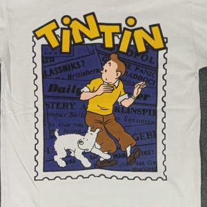 May include: White t-shirt featuring a graphic of Tintin, a cartoon character, and his dog Snowy. The design includes the word "TINTIN" in yellow, and a blue background with newspaper-style text. The shirt is made of a soft material.