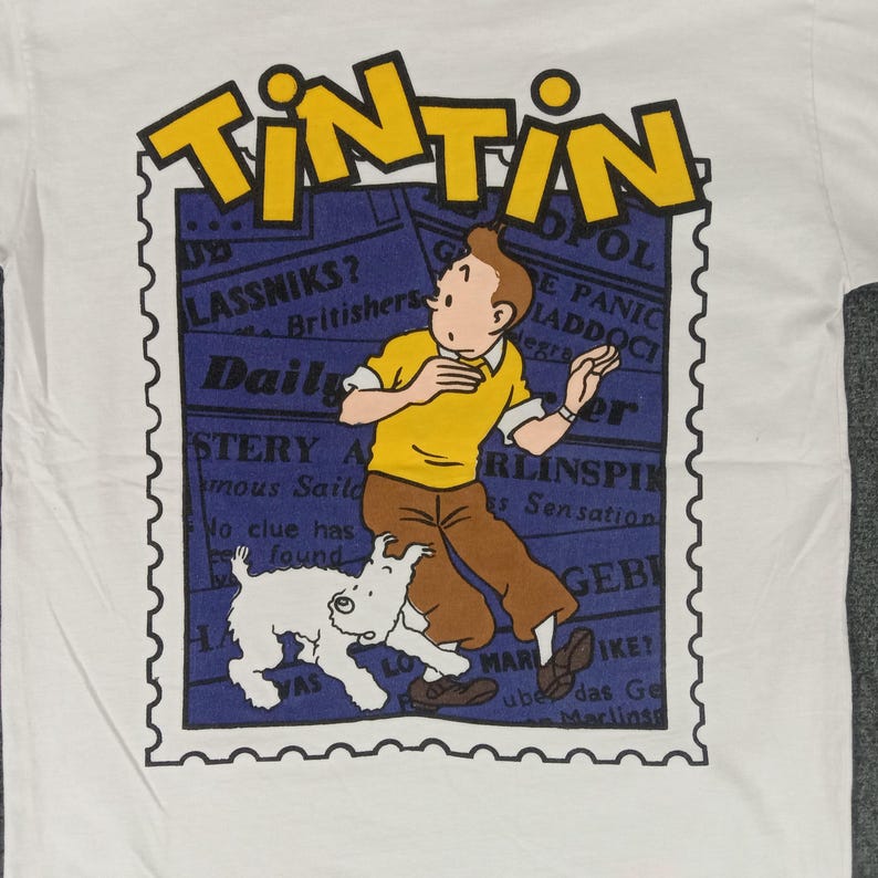 May include: White t-shirt featuring a graphic of Tintin, a cartoon character, and his dog Snowy. The design includes the word "TINTIN" in yellow, and a blue background with newspaper-style text. The shirt is made of a soft material.