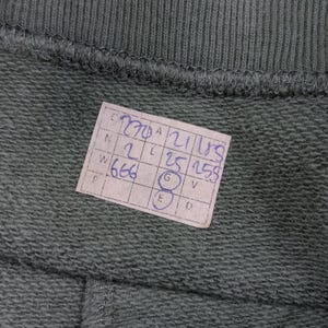 May include: Close-up of a section of olive green fabric, possibly a garment, with a stitched seam at the top and bottom. A small, rectangular tag with handwritten numbers and letters is attached to the fabric.