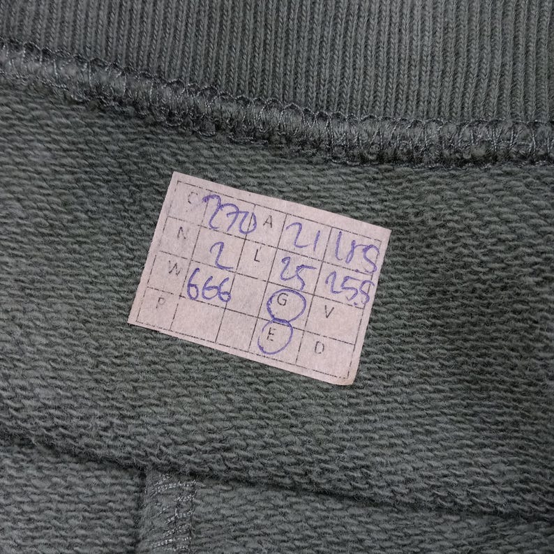 May include: Close-up of a section of olive green fabric, possibly a garment, with a stitched seam at the top and bottom. A small, rectangular tag with handwritten numbers and letters is attached to the fabric.