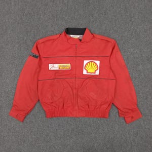 May include: Red racing jacket with black accents, featuring the Shell logo and other sponsor patches. The jacket has a black collar and a zipper closure. The jacket has two front pockets and a small pocket on the left sleeve.