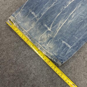Op de afbeelding: Close-up van een gedeelte van een blauwe jeans met witte, vervaagde markeringen. Een gele meetlint toont metingen in centimeters. De jeans ligt op een grijze ondergrond.