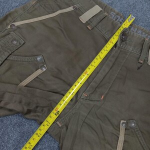 May include: Green cargo pants with a brown and beige trim. The pants have a zipper fly and button closure. The pants have a measuring tape on them, showing a measurement of 30 inches.