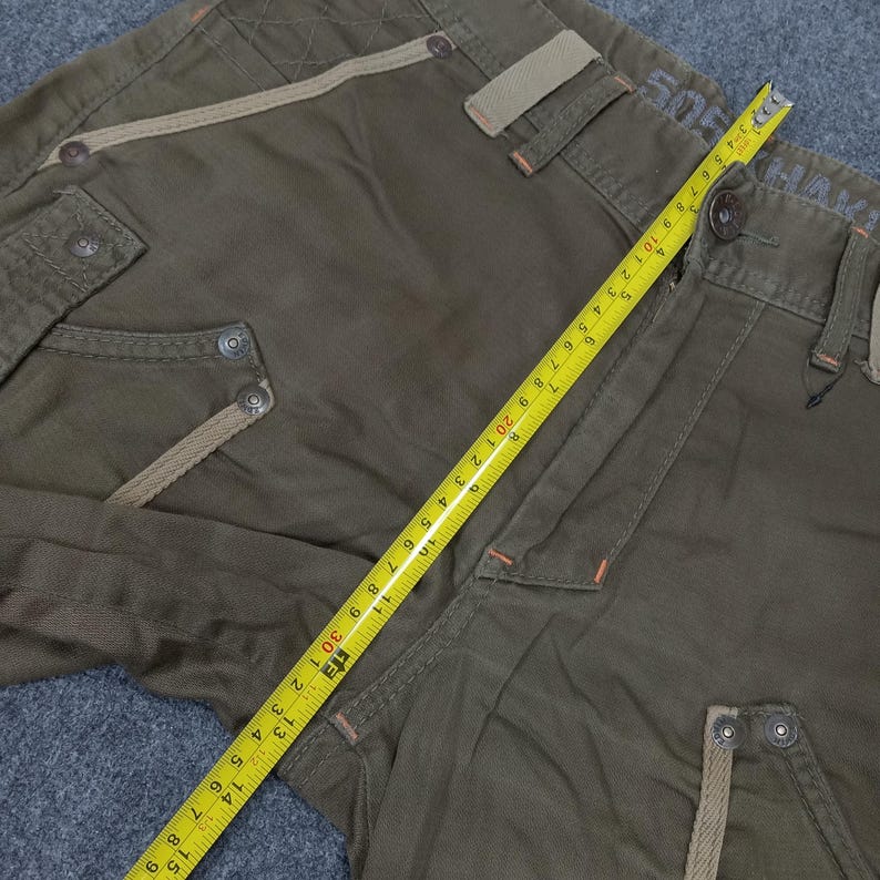 May include: Green cargo pants with a brown and beige trim. The pants have a zipper fly and button closure. The pants have a measuring tape on them, showing a measurement of 30 inches.