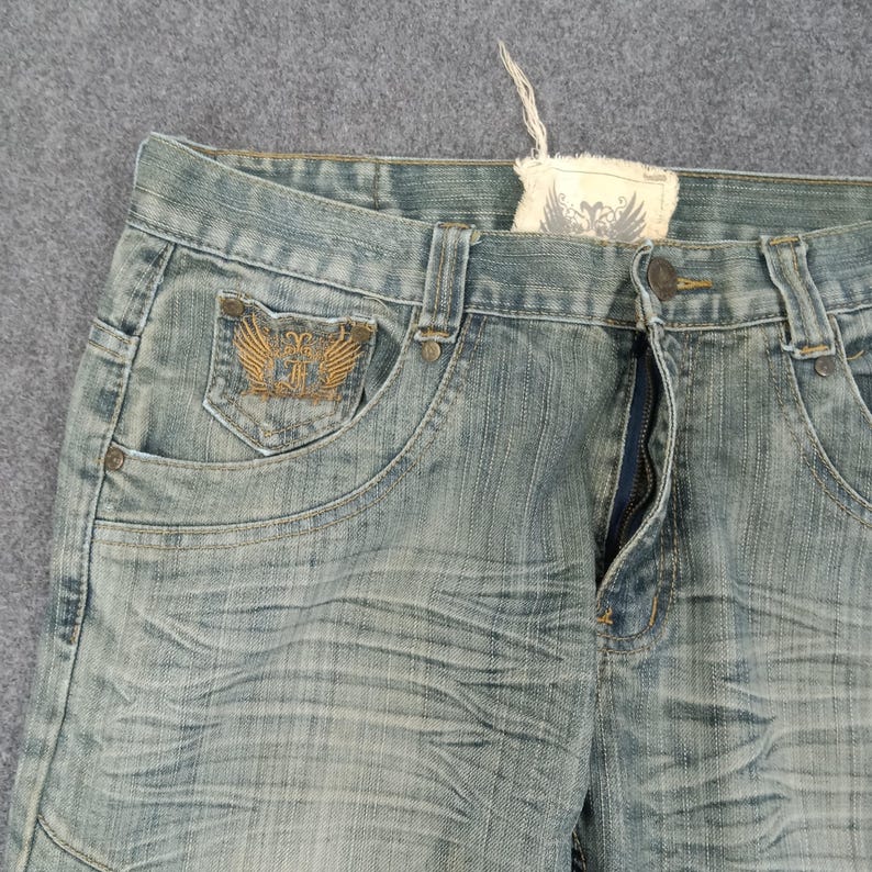 May include: Light blue denim jeans with a distressed wash. Features a decorative emblem on the front pocket and a partially unzipped fly. The jeans have a classic five-pocket design and a faded appearance.