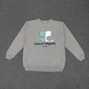 May include: A gray crew neck sweatshirt with the Courr&egrave;ges logo in teal, white, and navy blue. The logo features the brand name and "paris" in a sans-serif font. The sweatshirt is made of a soft material.