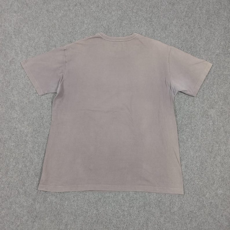 Puede incluir: Una camiseta lisa de manga corta, de color rosa empolvado. La camiseta de cuello redondo se muestra plana sobre una superficie gris, destacando su dise&ntilde;o sencillo y su estilo casual. La tela parece suave y ligeramente desgastada.