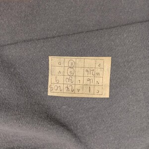 Puede incluir: Primer plano de una tela gris con una peque&ntilde;a etiqueta rectangular adjunta. La etiqueta tiene una cuadr&iacute;cula de n&uacute;meros y letras manuscritos, incluyendo "24 205" y "W216". La tela es de color gris liso y apagado.