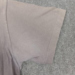 Puede incluir: Primer plano de una manga de camiseta gris. La manga es de un color gris liso y apagado. La tela parece suave y ligeramente texturizada. El borde de la manga est&aacute; cuidadosamente dobladillado.