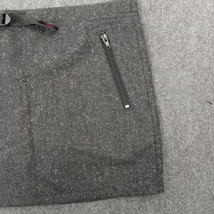 Pode incluir: Close-up de uma se&ccedil;&atilde;o de tecido cinza escuro com um padr&atilde;o salpicado. Um bolso com z&iacute;per preto &eacute; vis&iacute;vel no lado direito. Uma al&ccedil;a preta com fivela e detalhe vermelho est&aacute; no topo.