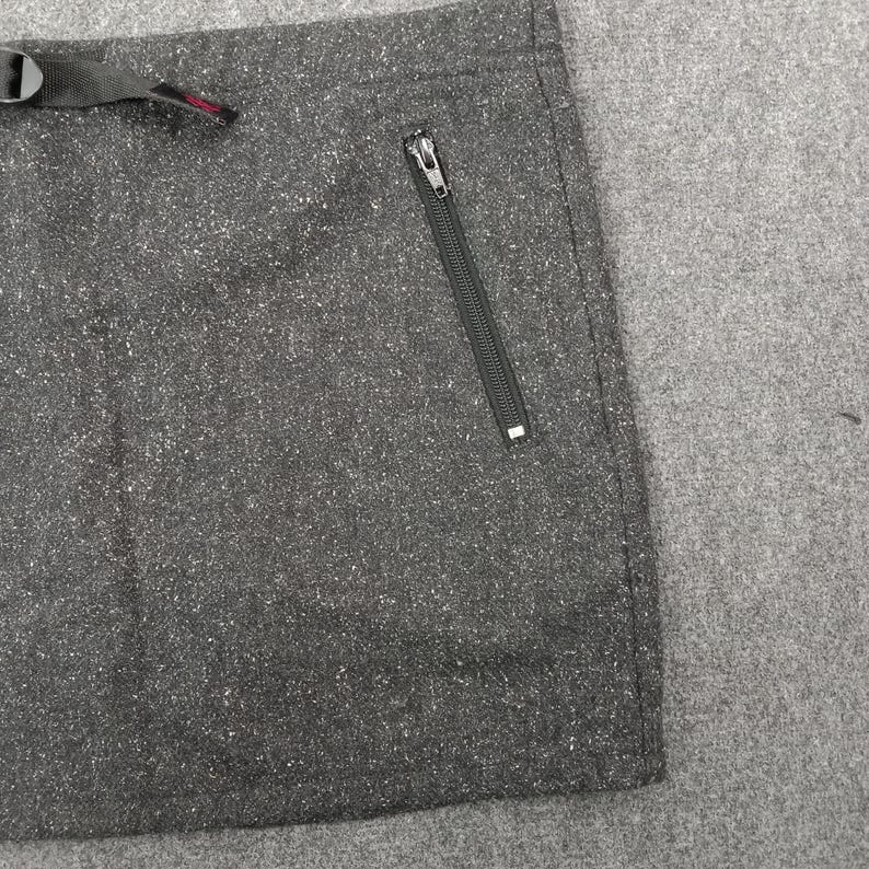 Pode incluir: Close-up de uma se&ccedil;&atilde;o de tecido cinza escuro com um padr&atilde;o salpicado. Um bolso com z&iacute;per preto &eacute; vis&iacute;vel no lado direito. Uma al&ccedil;a preta com fivela e detalhe vermelho est&aacute; no topo.