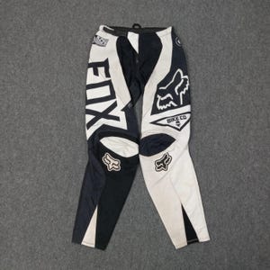 May include: Black and white motocross pants with the word "FOX" in large white letters on the left leg. The pants feature the Fox logo and "BIKE CO" text. The design includes black and white panels and a zipper closure.