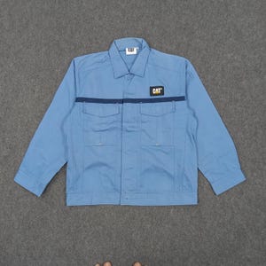 Puede incluir: Chaqueta de trabajo azul claro con el logotipo "CAT" en el pecho y el cuello. La chaqueta tiene dos bolsillos en el pecho y una franja azul oscuro en el pecho. La chaqueta de manga larga est&aacute; abotonada.