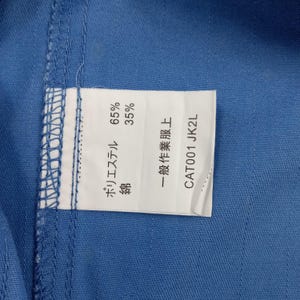 Puede incluir: Primer plano de una tela azul con una etiqueta blanca adjunta. La etiqueta muestra texto japon&eacute;s y porcentajes de poli&eacute;ster y algod&oacute;n. El texto "CAT001 JK2L" tambi&eacute;n es visible.