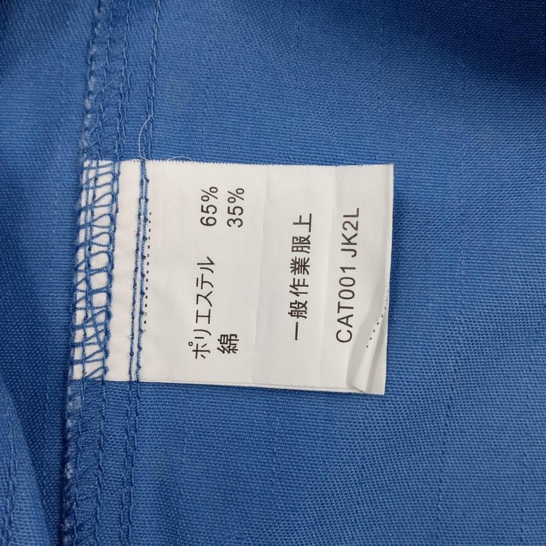 Puede incluir: Primer plano de una tela azul con una etiqueta blanca adjunta. La etiqueta muestra texto japon&eacute;s y porcentajes de poli&eacute;ster y algod&oacute;n. El texto "CAT001 JK2L" tambi&eacute;n es visible.