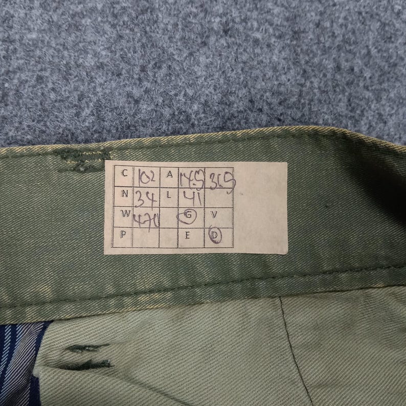 May include: A brown paper tag with handwritten measurements in a grid. The tag is attached to a green fabric garment. The measurements are: C 102, N 34, W 42, P, A 115, L 41, G, V, E, D.