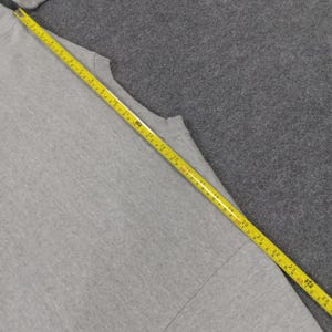 May include: A light gray t-shirt with a yellow measuring tape laid across it. The tape shows measurements in inches, with the shirt's length being measured. The shirt is on a gray surface.