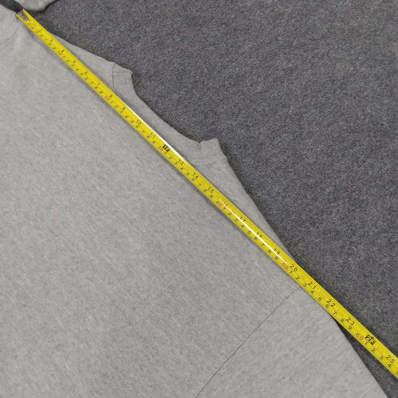 May include: A light gray t-shirt with a yellow measuring tape laid across it. The tape shows measurements in inches, with the shirt's length being measured. The shirt is on a gray surface.