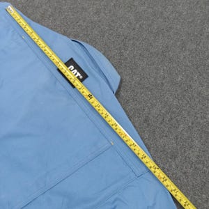 Puede incluir: Camisa de trabajo azul claro con un parche con el logo de CAT. Una cinta m&eacute;trica amarilla est&aacute; colocada sobre la camisa, mostrando medidas en cent&iacute;metros. La camisa tiene un bolsillo y costuras.