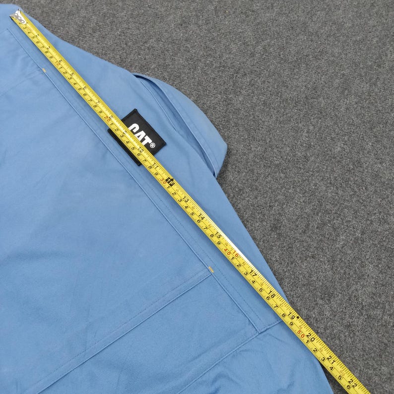 Puede incluir: Camisa de trabajo azul claro con un parche con el logo de CAT. Una cinta m&eacute;trica amarilla est&aacute; colocada sobre la camisa, mostrando medidas en cent&iacute;metros. La camisa tiene un bolsillo y costuras.