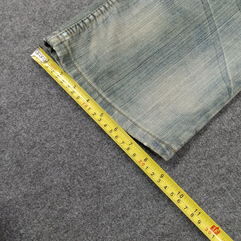 May include: Close-up of a section of light blue denim jeans. A yellow measuring tape is laid across the fabric, showing measurements in inches. The jeans have a faded appearance.