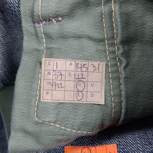 以下が含まれることがあります： デニムジーンズの内側のクローズアップ。手書きの数字と文字が書かれたタグが表示されています。タグは薄緑色の生地に付いています。ジーンズは青色のデニムです。