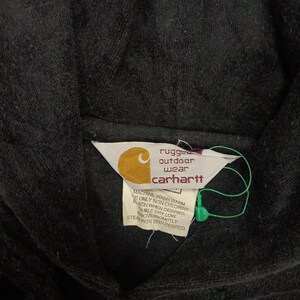 Puede incluir: Una etiqueta negra de Carhartt con texto blanco que dice "rugged outdoor wear Carhartt". La etiqueta tiene instrucciones para lavar y planchar la prenda.