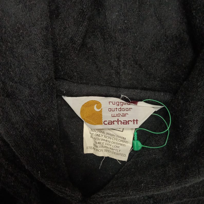 Puede incluir: Una etiqueta negra de Carhartt con texto blanco que dice "rugged outdoor wear Carhartt". La etiqueta tiene instrucciones para lavar y planchar la prenda.