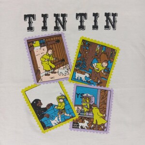 May include: White t-shirt featuring the word "TIN TIN" in black block letters above four colorful postage stamp-style illustrations. Each stamp depicts scenes with a boy and a white dog.