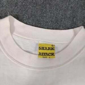 May include: White crew neck t-shirt with a yellow tag that reads "SHARK ATTACK" in black letters. The tag also includes the size "L". The shirt is made of a soft material.