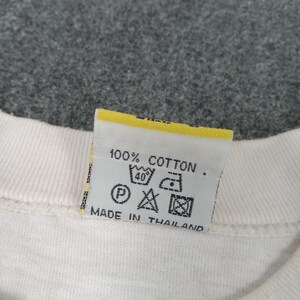 May include: Close-up of a white garment tag with yellow trim. The tag reads "100% Cotton" and includes washing instructions and "Made in Thailand". The background is a gray, textured fabric.