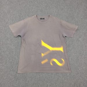 Puede incluir: Una camiseta gris de manga corta con un dise&ntilde;o amarillo de plantilla de las letras "Y2". La camiseta tiene cuello redondo y una peque&ntilde;a etiqueta cerca del cuello. La tela parece ligeramente descolorida.