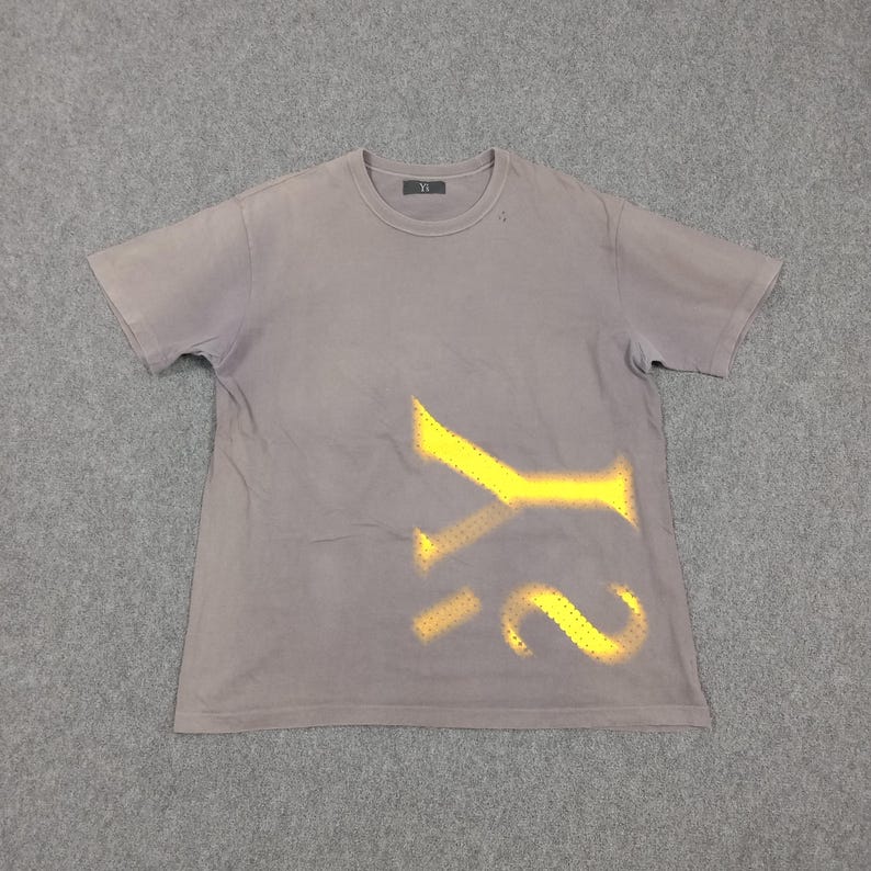 Puede incluir: Una camiseta gris de manga corta con un dise&ntilde;o amarillo de plantilla de las letras "Y2". La camiseta tiene cuello redondo y una peque&ntilde;a etiqueta cerca del cuello. La tela parece ligeramente descolorida.