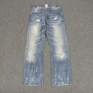 Op de afbeelding: Lichtblauwe spijkerbroek met distressed details, waaronder scheuren en vervaagde plekken. De jeans heeft een klassiek ontwerp met vijf zakken en een knoop- en ritssluiting. De denim heeft een versleten, vintage look.