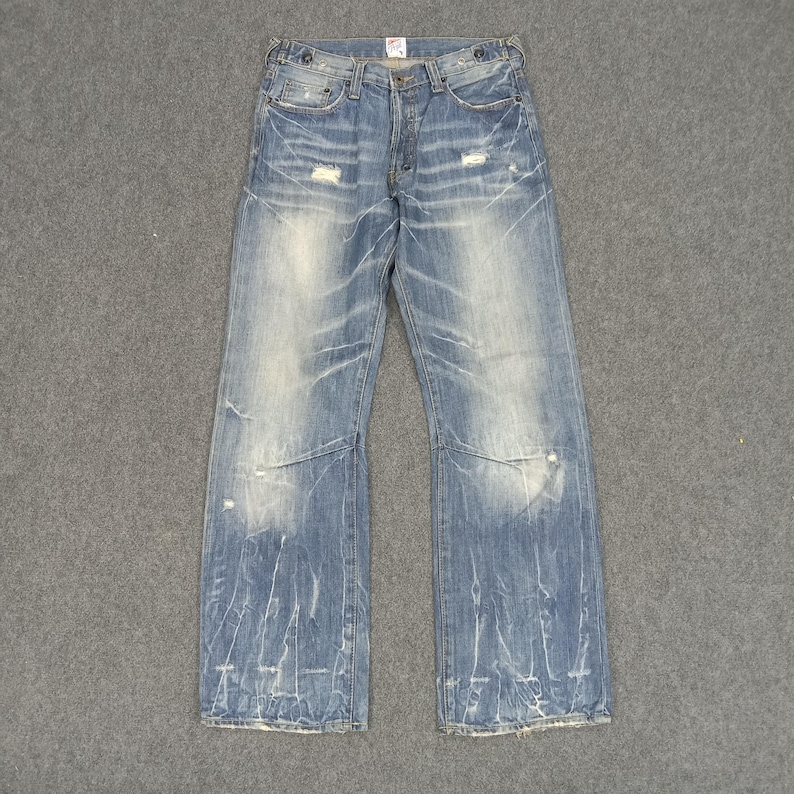 Op de afbeelding: Lichtblauwe spijkerbroek met distressed details, waaronder scheuren en vervaagde plekken. De jeans heeft een klassiek ontwerp met vijf zakken en een knoop- en ritssluiting. De denim heeft een versleten, vintage look.