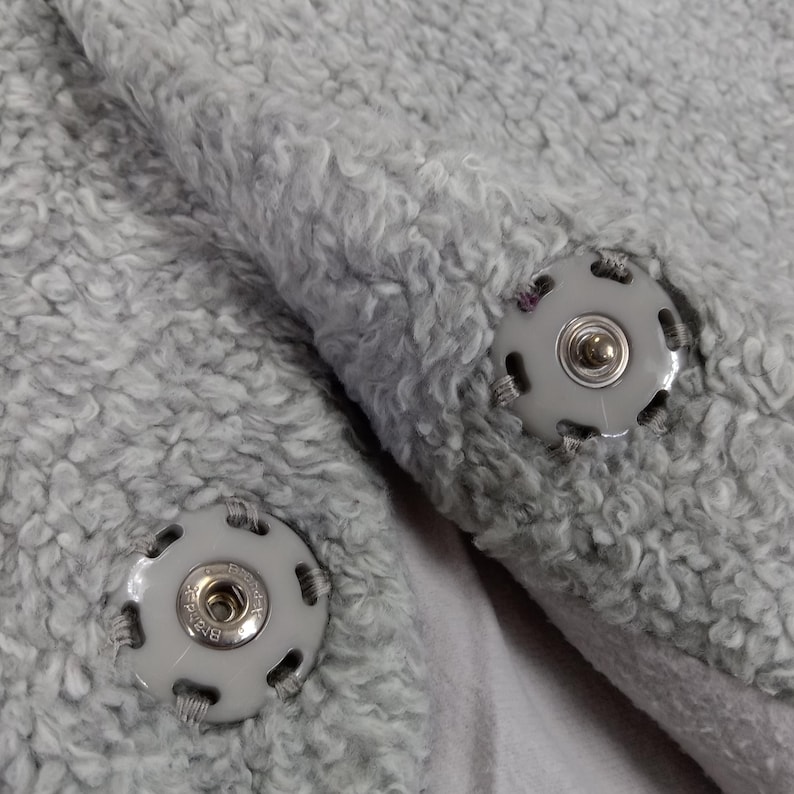 Op de afbeelding: Twee grijze stoffen sluitingen met een zilveren metalen knoop in het midden. De sluitingen zijn bevestigd aan een grijze, pluche stof.