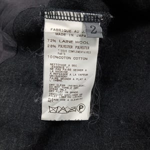 May include: A white garment care label attached to a black fabric surface. The label displays the text Made in Japan, size 2, and lists a composition of 72% wool and 28% polyester, with 100% cotton for complementary parts, plus care instructions.