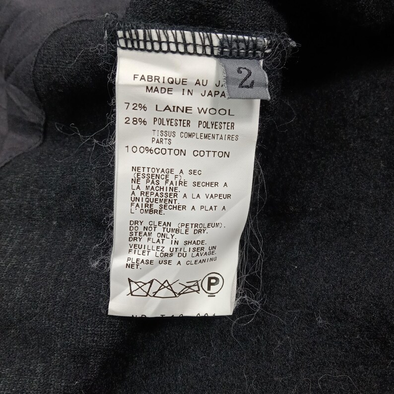 May include: A white garment care label attached to a black fabric surface. The label displays the text Made in Japan, size 2, and lists a composition of 72% wool and 28% polyester, with 100% cotton for complementary parts, plus care instructions.