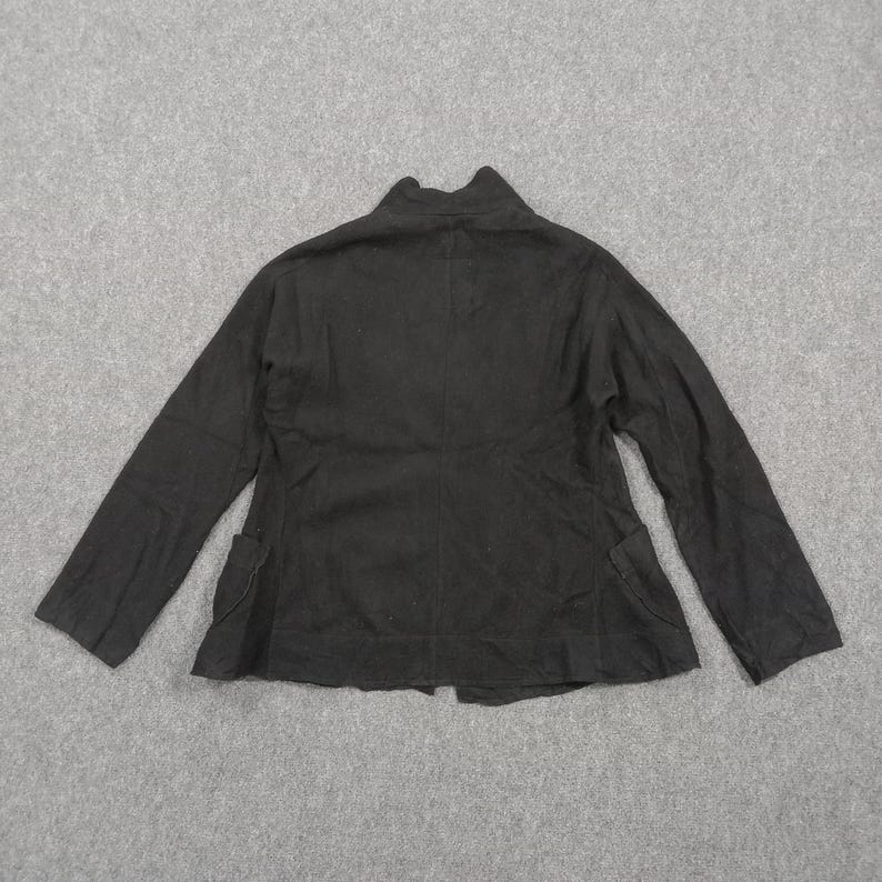 May include: A solid black long-sleeved jacket featuring a high collar, a central vertical seam down the back, and two patch pockets positioned at the lower front sides. The garment is constructed with a straight hemline and long sleeves.