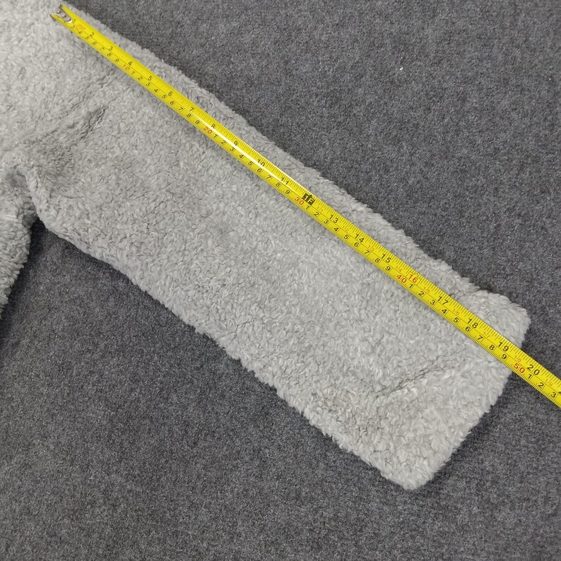 Op de afbeelding: Een close-up van een grijze, pluche stof met een gele meetlint erop. De tape meet tot 50 cm.