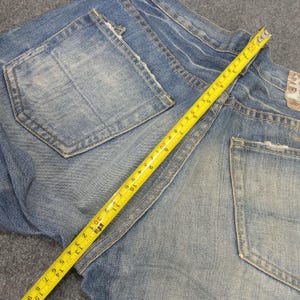 Op de afbeelding: Lichtblauwe spijkerbroek met distressed details, achterzakken en een gele meetlint die inches aangeeft. De jeans is opgevouwen, wat de textuur en slijtage van de stof laat zien.