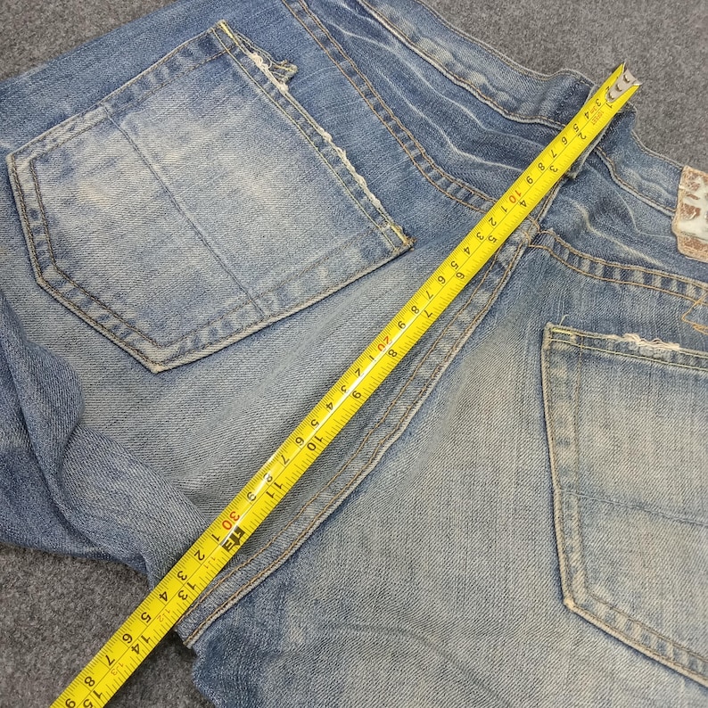 Op de afbeelding: Lichtblauwe spijkerbroek met distressed details, achterzakken en een gele meetlint die inches aangeeft. De jeans is opgevouwen, wat de textuur en slijtage van de stof laat zien.