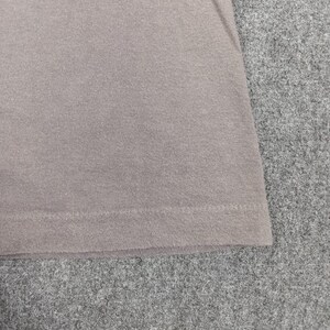 Puede incluir: Primer plano de una secci&oacute;n de una camiseta marr&oacute;n claro. La tela tiene un aspecto texturizado, con l&iacute;neas de punto visibles. El dobladillo inferior es visible, mostrando un borde doblado y cosido. La camiseta est&aacute; sobre una superficie gris.