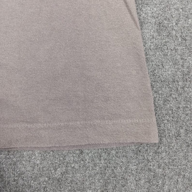 Puede incluir: Primer plano de una secci&oacute;n de una camiseta marr&oacute;n claro. La tela tiene un aspecto texturizado, con l&iacute;neas de punto visibles. El dobladillo inferior es visible, mostrando un borde doblado y cosido. La camiseta est&aacute; sobre una superficie gris.